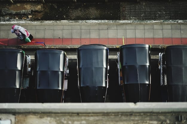 Several large, dark-colored rainwater collection barrels are neatly lined up along the side of a residential building. The barrels are connected to the downspouts of the building's gutters and have spigots for easy access to the collected water. The area around the barrels is clean and well-maintained.